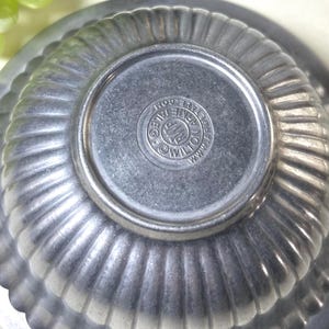 Op de afbeelding: Close-up van een vintage, gegroefde metalen kom met een cirkelvormige basis. De kom heeft een geribbeld ontwerp en een gestempeld logo op de bodem. Het logo luidt "Wilton Armetale Co."