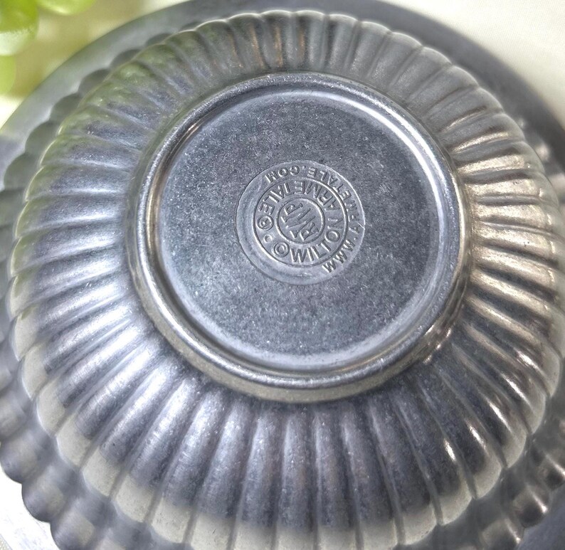 Op de afbeelding: Close-up van een vintage, gegroefde metalen kom met een cirkelvormige basis. De kom heeft een geribbeld ontwerp en een gestempeld logo op de bodem. Het logo luidt "Wilton Armetale Co."