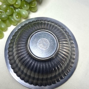 Op de afbeelding: Een tinnen, gegroefde kom met een cirkelvormige basis. De kom heeft een geschulpte rand en een cirkelvormig embleem in het midden. De kom is waarschijnlijk vintage en geschikt om te serveren of tentoon te stellen. De kom wordt van onderaf getoond.