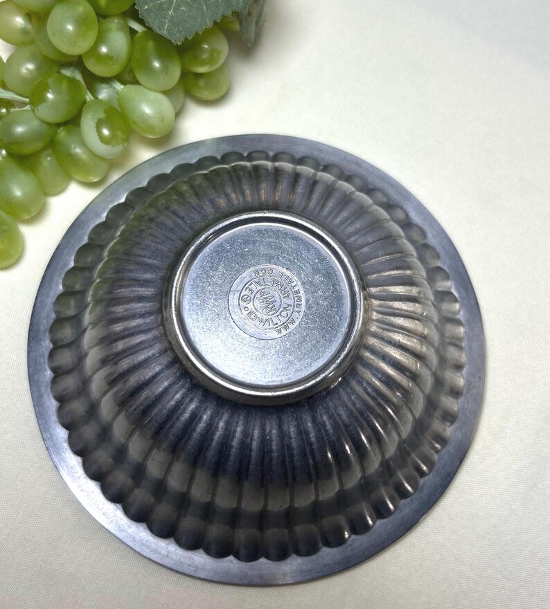 Op de afbeelding: Een tinnen, gegroefde kom met een cirkelvormige basis. De kom heeft een geschulpte rand en een cirkelvormig embleem in het midden. De kom is waarschijnlijk vintage en geschikt om te serveren of tentoon te stellen. De kom wordt van onderaf getoond.