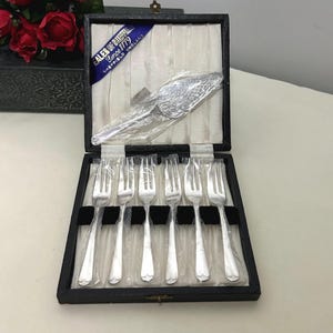 May include: A silver-plated cake server and six matching forks presented in a black hinged box with a cream-colored satin lining. The blue label inside reads Eales of Sheffield Since 1779 Sheffield England. Each piece is wrapped in clear plastic.