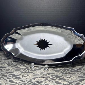 May include: An oval silver-toned metal serving tray with a scalloped edge and a reflective finish. The center of the tray features a prominent, dark, multi-pointed starburst or sunburst graphic design that contrasts with the polished surface.