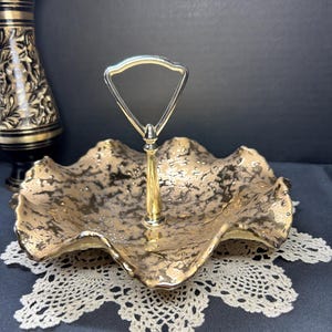 May include: A gold-toned, two-tiered serving tray with a wavy rim and a handle. The tray has a textured surface with dark accents. It sits on a white lace doily. The handle is a silver-colored metal.