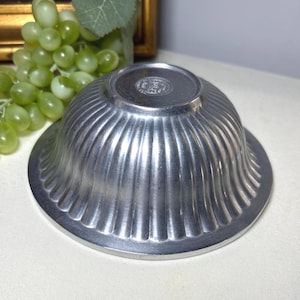 Op de afbeelding: Een kleine, zilverkleurige kom met een gegroefd ontwerp. De kom heeft een brede, platte basis en een afgeronde bovenkant. De kom is gemaakt van metaal en heeft een glanzende afwerking. De kom is waarschijnlijk vintage of antiek.