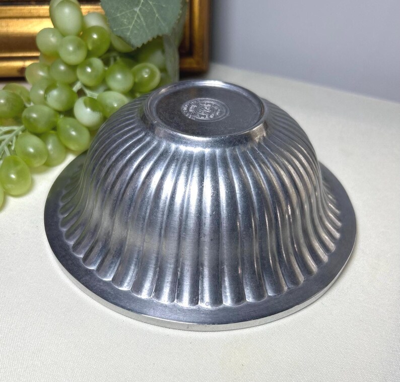 Op de afbeelding: Een kleine, zilverkleurige kom met een gegroefd ontwerp. De kom heeft een brede, platte basis en een afgeronde bovenkant. De kom is gemaakt van metaal en heeft een glanzende afwerking. De kom is waarschijnlijk vintage of antiek.