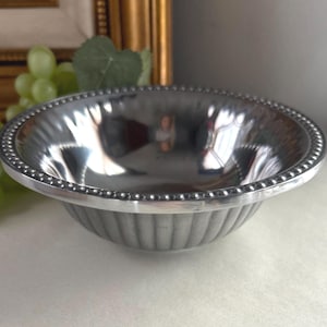 Op de afbeelding: Een kleine, zilverkleurige kom met een kralenrand en verticaal geribbelde zijkanten. De kom reflecteert de omgeving en cre&euml;ert een spiegeleffect. De kom is waarschijnlijk gemaakt van metaal en is geschikt om te serveren of tentoon te stellen.