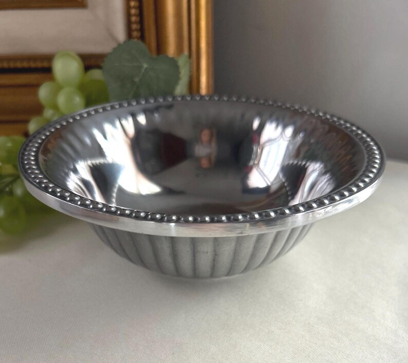 Op de afbeelding: Een kleine, zilverkleurige kom met een kralenrand en verticaal geribbelde zijkanten. De kom reflecteert de omgeving en cre&euml;ert een spiegeleffect. De kom is waarschijnlijk gemaakt van metaal en is geschikt om te serveren of tentoon te stellen.