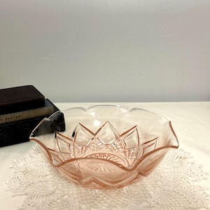 May include: A pale pink glass bowl featuring a scalloped rim and a geometric starburst pattern pressed into the interior base and sides. The bowl has a translucent appearance with faceted edges that catch the light, creating a clear pink hue.