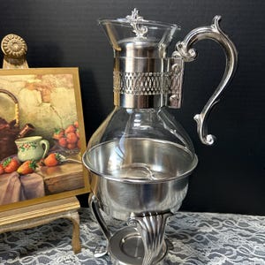 May include: A silver-toned, vintage tea or coffee server with a clear glass carafe. The server has an ornate handle and decorative accents. The base has a burner area and is supported by three decorative legs. A small painting is in the background.