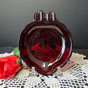 May include: A ruby red glass dish in a leaf shape. The dish has a scalloped edge and sits on two clear plastic stands. A red rose is visible in the background. The dish is likely vintage.