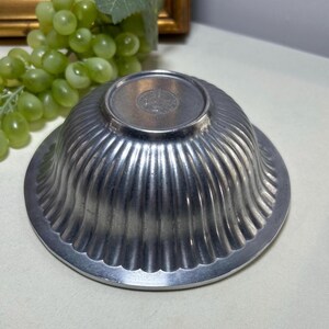 Op de afbeelding: Een zilverkleurige, gegroefde kom met een geschulpte rand. De kom staat ondersteboven en onthult de basis en het ingewikkelde ontwerp van de gegroefde buitenkant. De kom is waarschijnlijk gemaakt van metaal en heeft een vintage of antieke uitstraling.