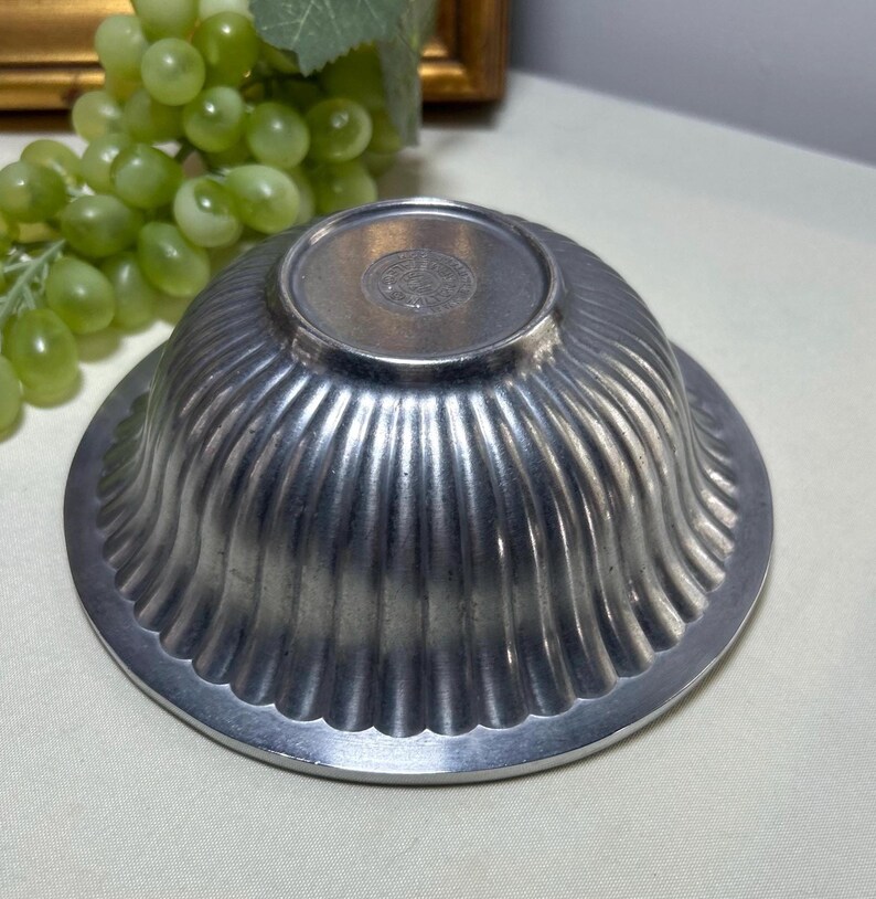 Op de afbeelding: Een zilverkleurige, gegroefde kom met een geschulpte rand. De kom staat ondersteboven en onthult de basis en het ingewikkelde ontwerp van de gegroefde buitenkant. De kom is waarschijnlijk gemaakt van metaal en heeft een vintage of antieke uitstraling.