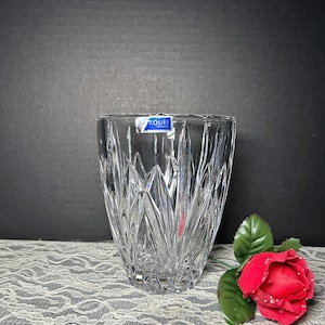 May include: Clear crystal vase with a textured, leaf-like pattern. The vase has a flared shape and a blue and white label that reads "MARQUIS". A red rose with green leaves rests nearby on a white lace surface.
