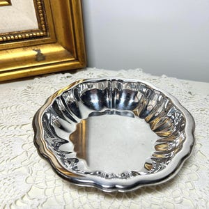May include: A small, silver-colored dish with a scalloped edge. The dish has a reflective surface and a shallow bowl shape. The dish is likely made of metal and is suitable for serving small portions of food or holding jewelry.