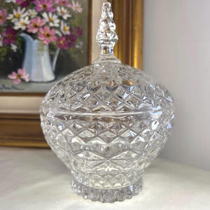 May include: A clear, diamond-cut glass candy dish with a lid. The dish has a round body and a pedestal base. The lid is topped with a decorative finial. The dish is likely vintage and sits on a white surface.