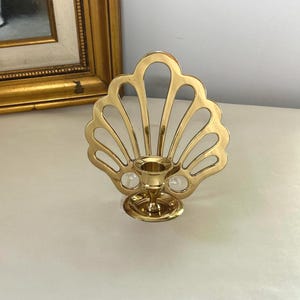 May include: A polished gold-tone brass candle holder featuring a decorative fan-shaped backplate with a scalloped edge and openwork cutouts. The central circular base supports a small, cylindrical candle cup designed to hold a single taper candle.