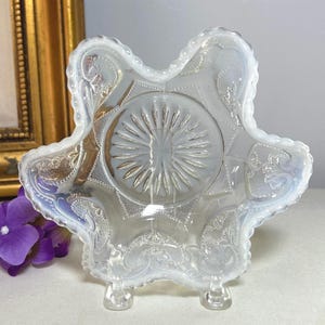 May include: A clear, opalescent glass bowl with a starburst design in the center and ornate, embossed patterns. The bowl has a scalloped edge and sits on three small feet. The bowl is likely vintage or antique.