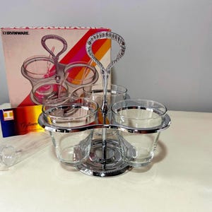 May include: A chrome-plated condiment server set featuring three clear glass bowls held in a circular metal frame with a central carrying handle. The set includes a box labeled IRVINWARE with a graphic illustration of the product on the front.