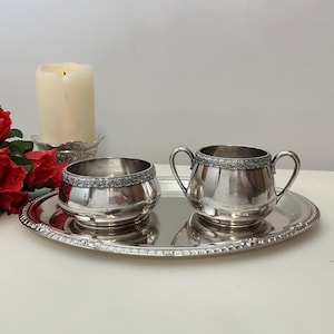 May include: A silver-toned metal serving set consisting of an oval tray, a small open bowl, and a two-handled sugar bowl. Each piece features a decorative embossed floral border along the rim. The set is arranged on a flat surface.