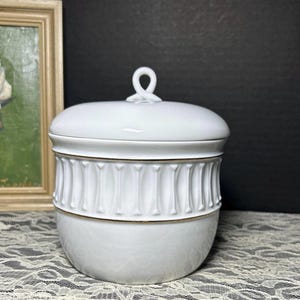 May include: White ceramic lidded jar with a decorative band of vertical lines and a gold trim. The lid has a loop handle. The jar is sitting on a white lace surface.