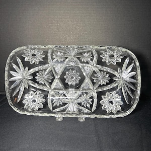 May include: A clear, rectangular glass serving dish with a starburst and floral pattern. The dish has a slightly raised base and is designed with intricate geometric shapes and star-like designs. The edges are rounded.