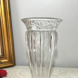 May include: A clear, elegant glass vase with a flared top and vertical ribbed design. The rim features a decorative leaf pattern. The vase is displayed on a white lace tablecloth, with a gold framed picture in the background.