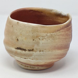 May include: A small, hand-thrown ceramic bowl with a light brown glaze and a slightly uneven rim. The bowl has a subtle, swirling pattern in the glaze, creating a unique and rustic look.