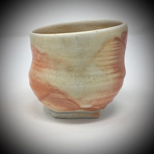 May include: A small, off-white ceramic cup with a square base and a slightly flared rim. The cup has a mottled glaze with shades of brown and beige.