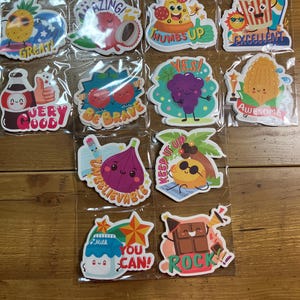 Puede incluir: Una colección de pegatinas coloridas con varios alimentos y mensajes positivos. Los diseños incluyen una piña, pizza, palomitas de maíz, uvas, maíz y una botella. Textos incluyen "Great!", "Thumbs Up", "Excellent" y "Very Good".