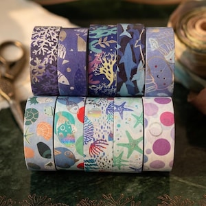 May include: A collection of eight decorative washi tape rolls. The tapes feature various ocean-themed designs, including coral, fish, starfish, and jellyfish, in shades of blue, purple, and green. Some rolls have metallic gold accents. The tapes are arranged on a dark green surface.