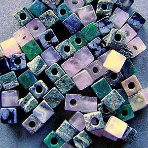 Puede incluir: Una colección de pequeñas cuentas de forma cuadrada en varios colores y texturas. Las cuentas presentan un orificio central y están hechas de diferentes tipos de piedra, incluyendo tonos de verde, azul, azul marino oscuro y rosa pálido o blanco.