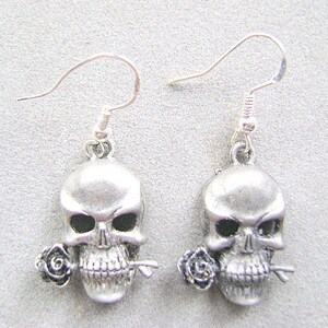 May include: A pair of silver skull earrings with a single black rose on each skull. The earrings have a simple hook closure.