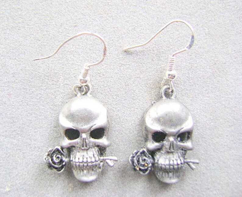 May include: A pair of silver skull earrings with a single black rose on each skull. The earrings have a simple hook closure.