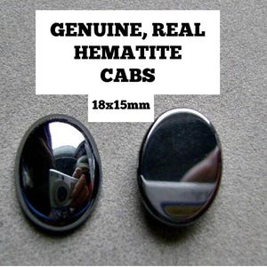 May include: Two oval-shaped, polished hematite cabochons with a metallic, mirror-like finish. The text above the stones reads: GENUINE, REAL HEMATITE CABS 18x15mm. These dark gray, smooth stones feature a reflective surface that shows a clear reflection.
