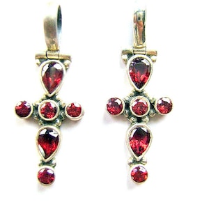May include: Two silver-toned cross pendants with teardrop and round, deep red gemstones. The crosses have a loop at the top for hanging. The gemstones are set in a silver metal setting. The background is white.