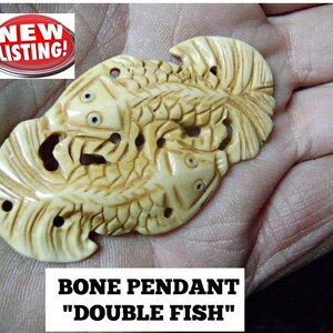 以下が含まれることがあります： 2匹の魚が絡み合った、彫刻された骨のペンダント。ペンダントは薄いベージュ色で、細部までこだわったデザインが施されています。ペンダントの下には「BONE PENDANT "DOUBLE FISH"」と書いてあります。