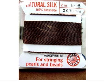 Puede incluir: Un carrete de hilo de seda natural marrón oscuro para ensartar perlas y cuentas. El empaque muestra el texto NATURAL SILK, 2 m, No. 6, 0,70 mm y www.griffin.de. El nombre de la marca Griffin está impreso en un logo circular rojo.