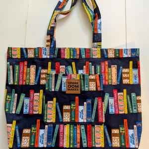 May include: A navy blue tote bag featuring a colorful bookshelf design. The bag displays various book titles and spines in a vertical arrangement. The handles and a small patch with the text "SPARE OOM MAKES" are also visible.