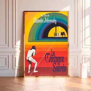 May include: A vintage movie poster for the film "La Montagne Sacrée" directed by Alejandro Jodorowsky. The poster features a colorful rainbow archway and a man in a loincloth holding a bow and arrow. The text "La Montagne Sacrée" is in blue letters on a red background.
