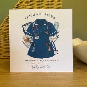 May include: A square greeting card featuring a dark blue nurse uniform top surrounded by medical illustrations including a stethoscope, clipboard, coffee cup, and thermometer. The text reads CONGRATULATIONS ON BECOMING A QUALIFIED NURSE and Olivia.