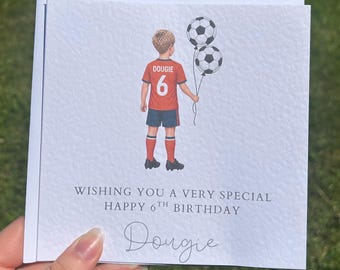 以下が含まれることがあります： 背中にDougieという名前と背番号6が入った赤いサッカーユニフォームを着た子供のイラストが描かれた白いグリーティングカード。子供は2つのサッカーボールの風船を持っています。テキストには「WISHING YOU A VERY SPECIAL HAPPY 6TH BIRTHDAY」と書かれています。