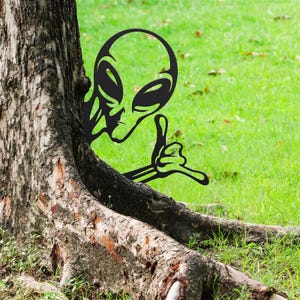 May include: Black metal alien silhouette yard art. The cutout depicts an alien head with large eyes and a hand giving a thumbs-up gesture. The art is placed near a tree trunk on a grassy lawn.