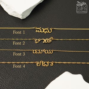 May include: Four gold-toned necklaces featuring custom name pendants in Kannada script, each displayed with a different chain style and font labeled Font 1, Font 2, Font 3, and Font 4. The jewelry is arranged horizontally against a black textured background.