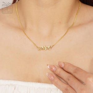 May include: A gold-colored necklace featuring a delicate chain and a central pendant displaying the Thai script word "อาทิตย์". The metal has a polished, reflective finish, and the pendant is integrated directly into the chain links.