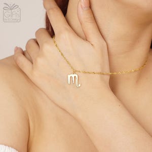 May include: A gold-toned necklace featuring a delicate rope chain and a pendant shaped like the Scorpio zodiac symbol. The pendant is a polished gold-colored metal cutout of the letter M with a curved tail ending in an arrow point.
