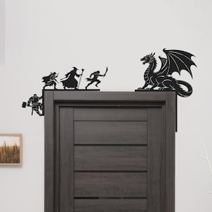 May include: Black metal wall art featuring a dragon and several fantasy figures. The dragon is perched on a corner, while the figures appear to be running. The figures include a warrior, a wizard, and a swordsman. The art is mounted on a dark brown door.