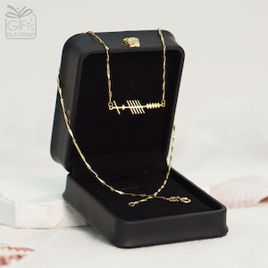 May include: A gold-colored necklace with a unique design, presented in an open black jewelry box. The necklace features a series of vertical lines and a horizontal line with a small cross-like shape. The box has a small, decorative gold accent on the lid.