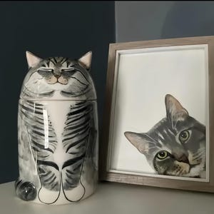 May include: A ceramic cat-shaped jar with a lid, painted in gray and white stripes, sits next to a framed watercolor painting of a cat's face. The jar has a smiling cat face. The painting is in a light wood frame.