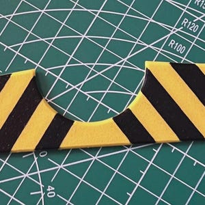May include: A yellow and black striped object, possibly a craft or construction component, lies on a green cutting mat. The object has a unique shape with a curved cutout in the center and diagonal stripes.