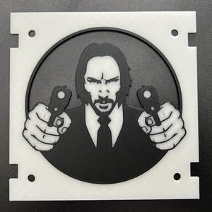 May include: A black and white 3D-printed image of a man holding two guns. The image is set within a circular black frame, surrounded by a white square with cut-out corners and four small holes.